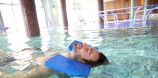 A 30-something year old man floats in the pool on his back with a floating pillow supporting his head.