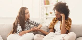 Two friends sit on the couch. One is a white woman and is supportively holding the hand of her black roommate who has some sort of head pain.